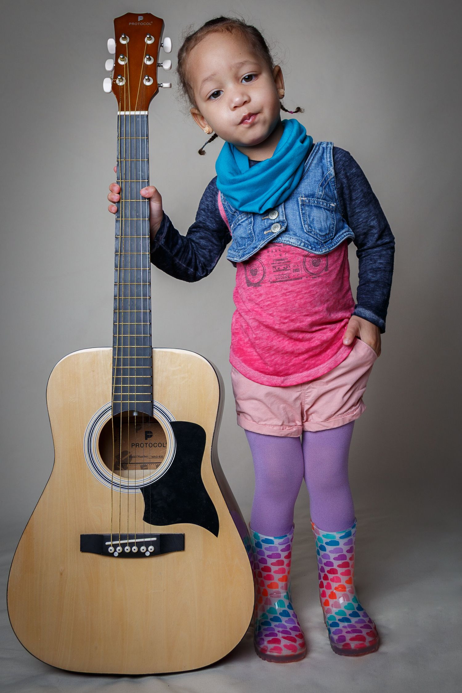 A small child in colorful socks and denim jacket poses playfully with an acoustic guitar against a gray studio backdrop.