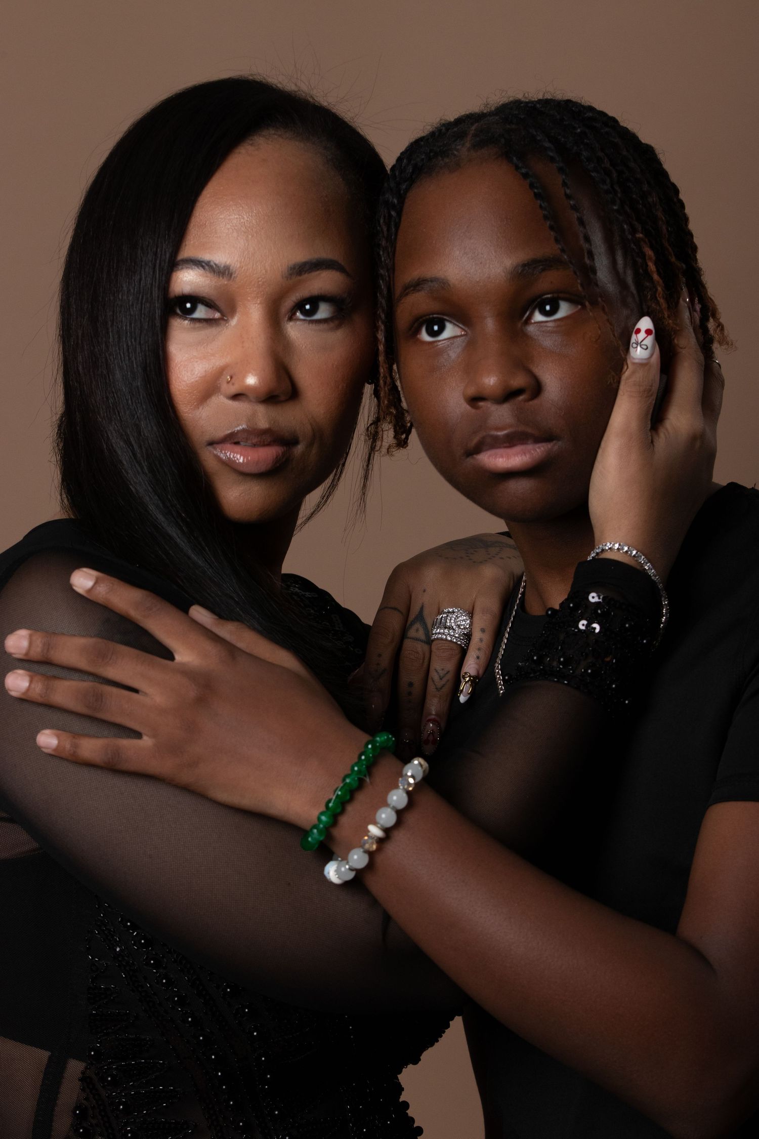 Two Black women in black outfits pose together, wearing bracelets and rings, against a warm brown background.