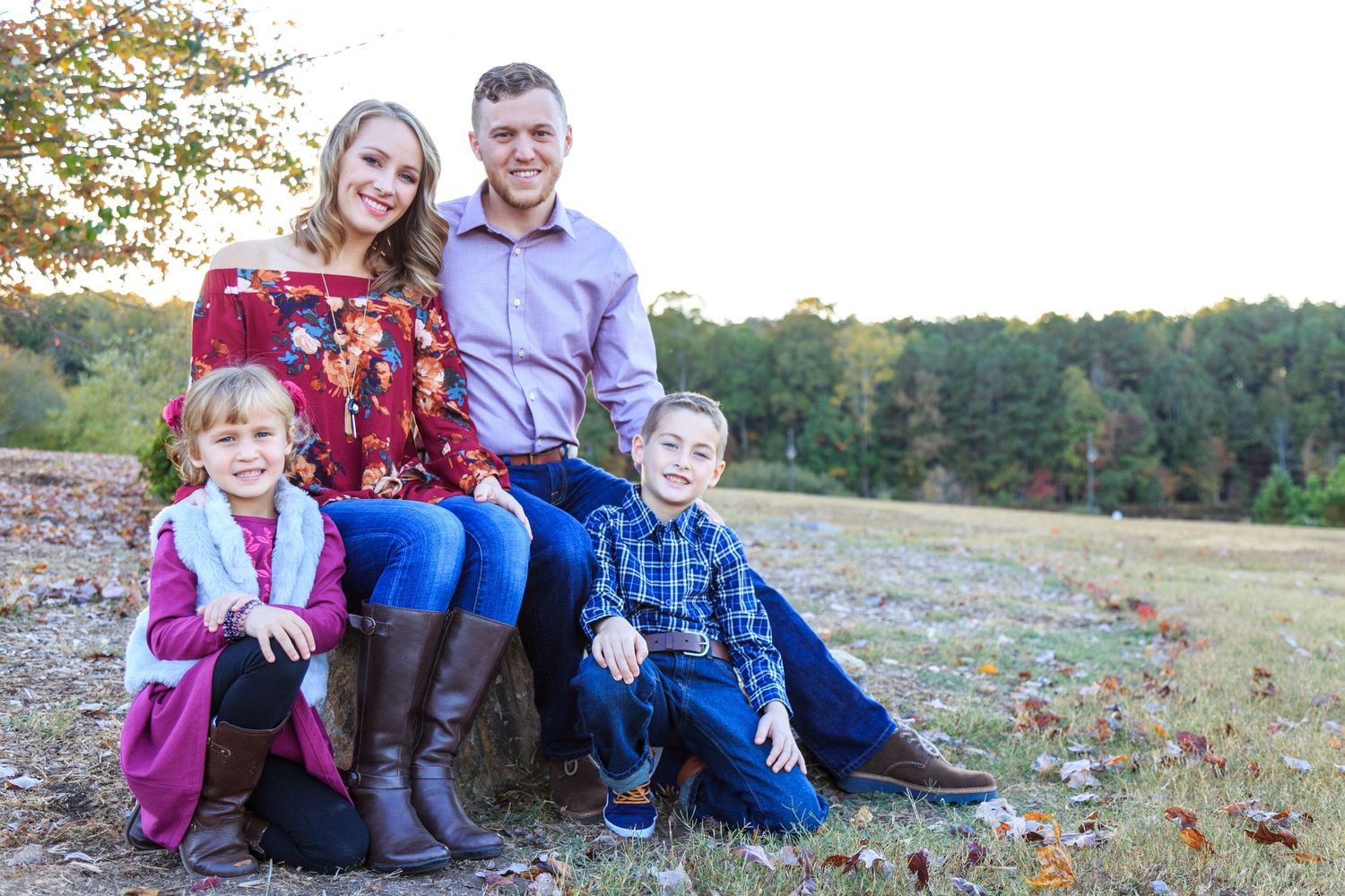 Family sitting together on autumn grass with colorful fall leaves scattered around during golden hour sunset.