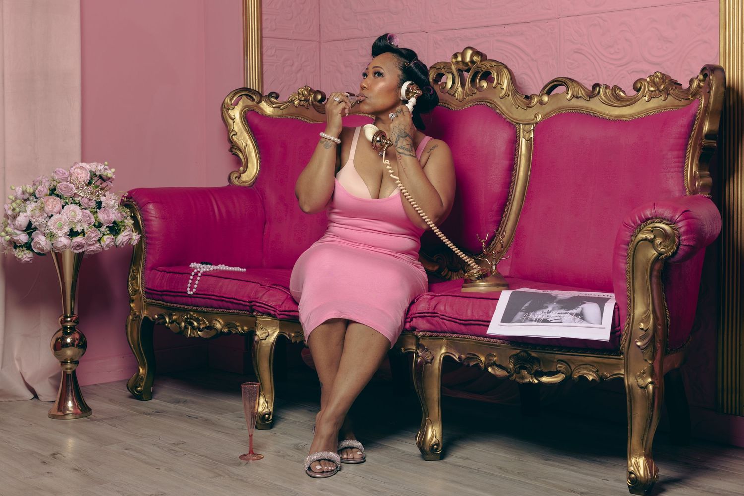 Woman in pink dress sitting on ornate gold and pink sofa, talking on a retro phone in a glamorous pink room.