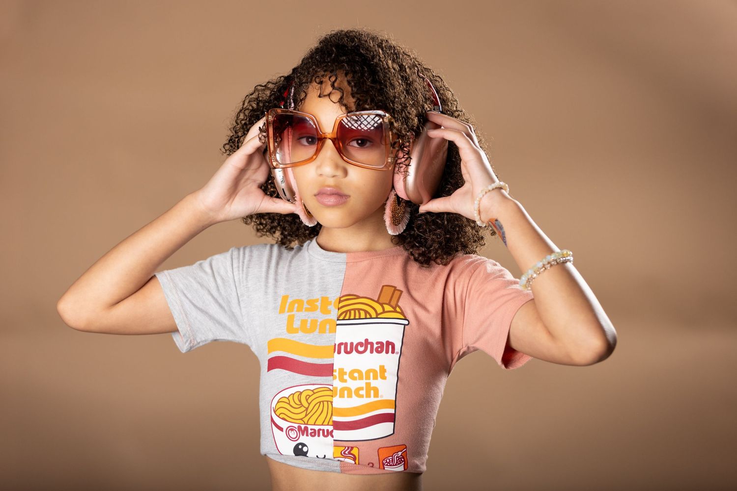 A person wearing sunglasses and a graphic t-shirt poses with curly hair against a brown background.