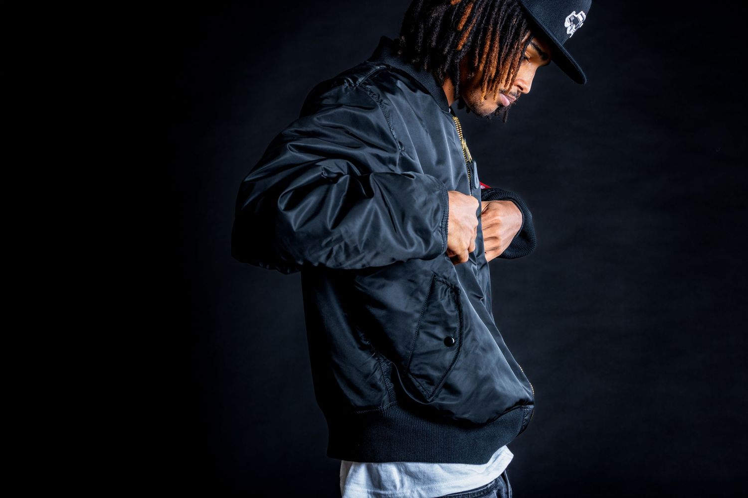 A person wearing a black windbreaker jacket and baseball cap poses against a dark background.