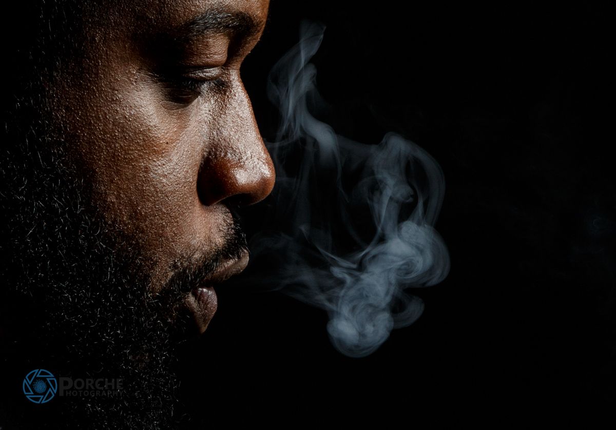 A dark silhouette exhales smoke against a black background in a moody artistic portrait.