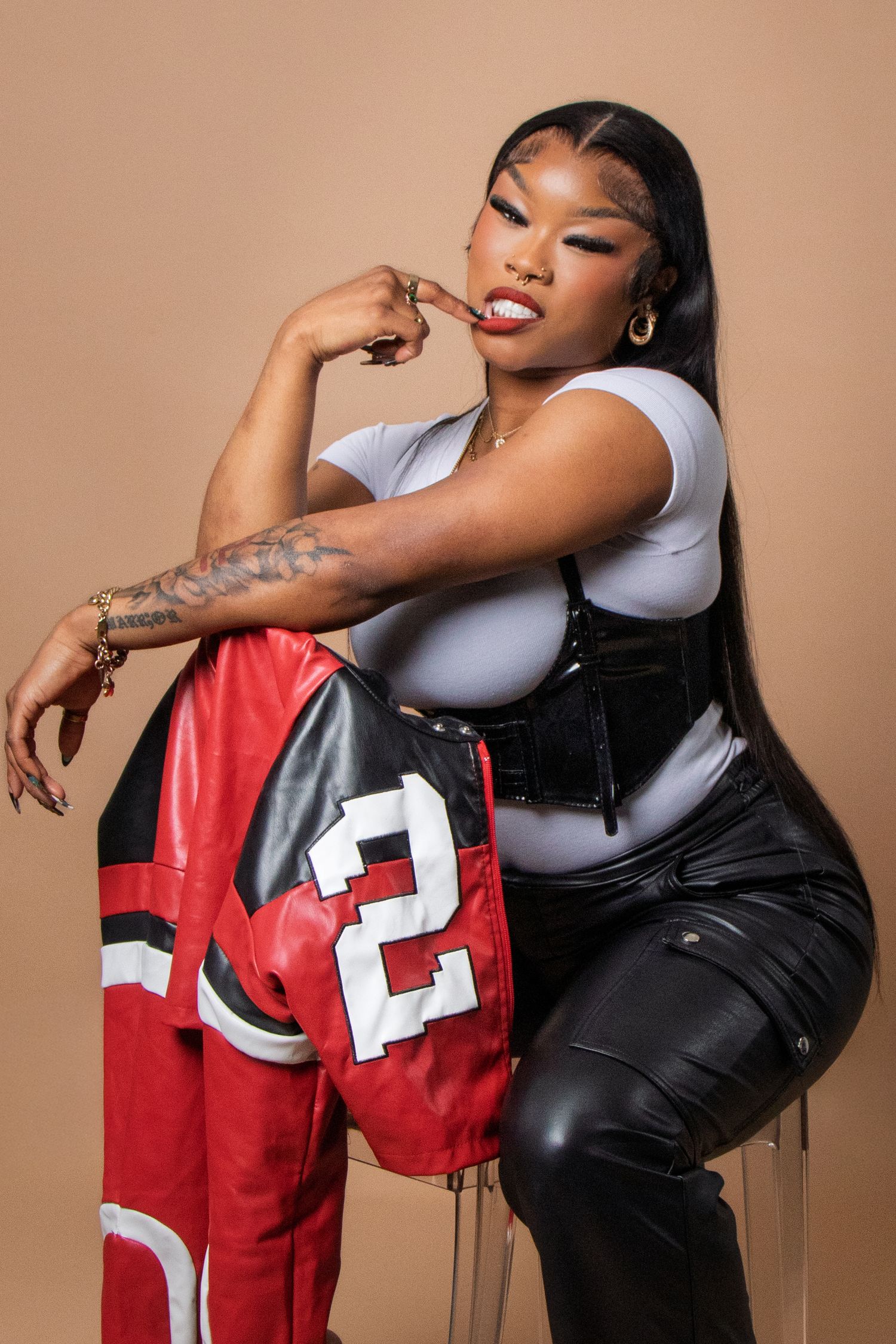 Woman in white bodysuit, black corset, and leather pants poses with red varsity jacket in studio.