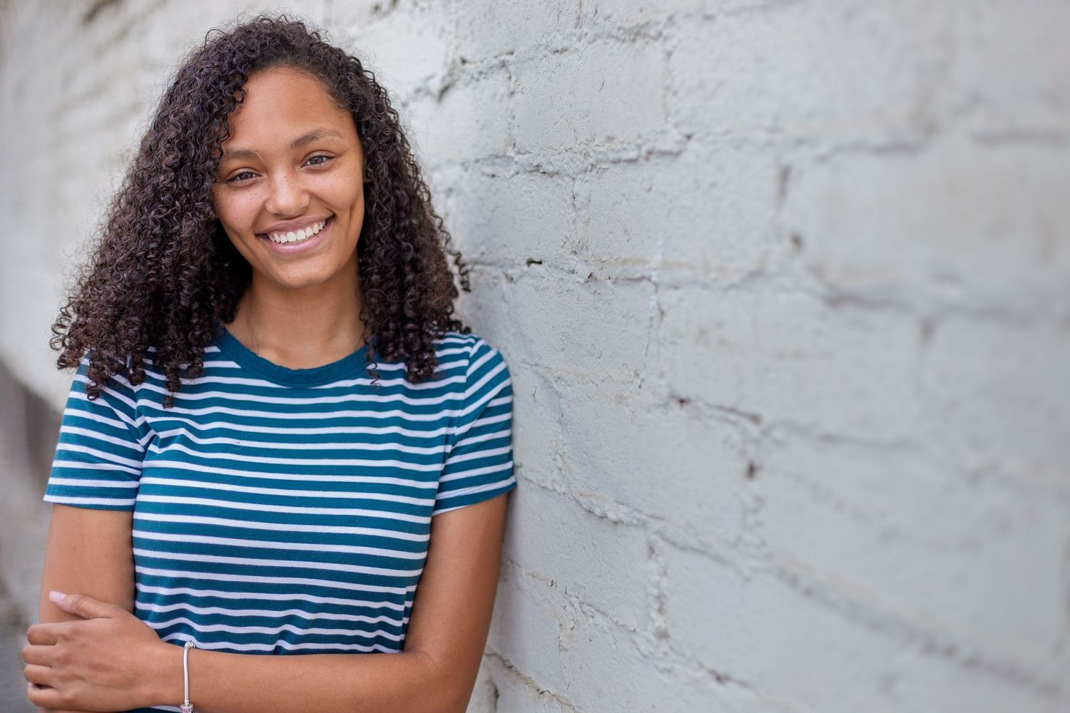 Person with curly hair wearing a blue and white striped shirt smiles against a gray wall.