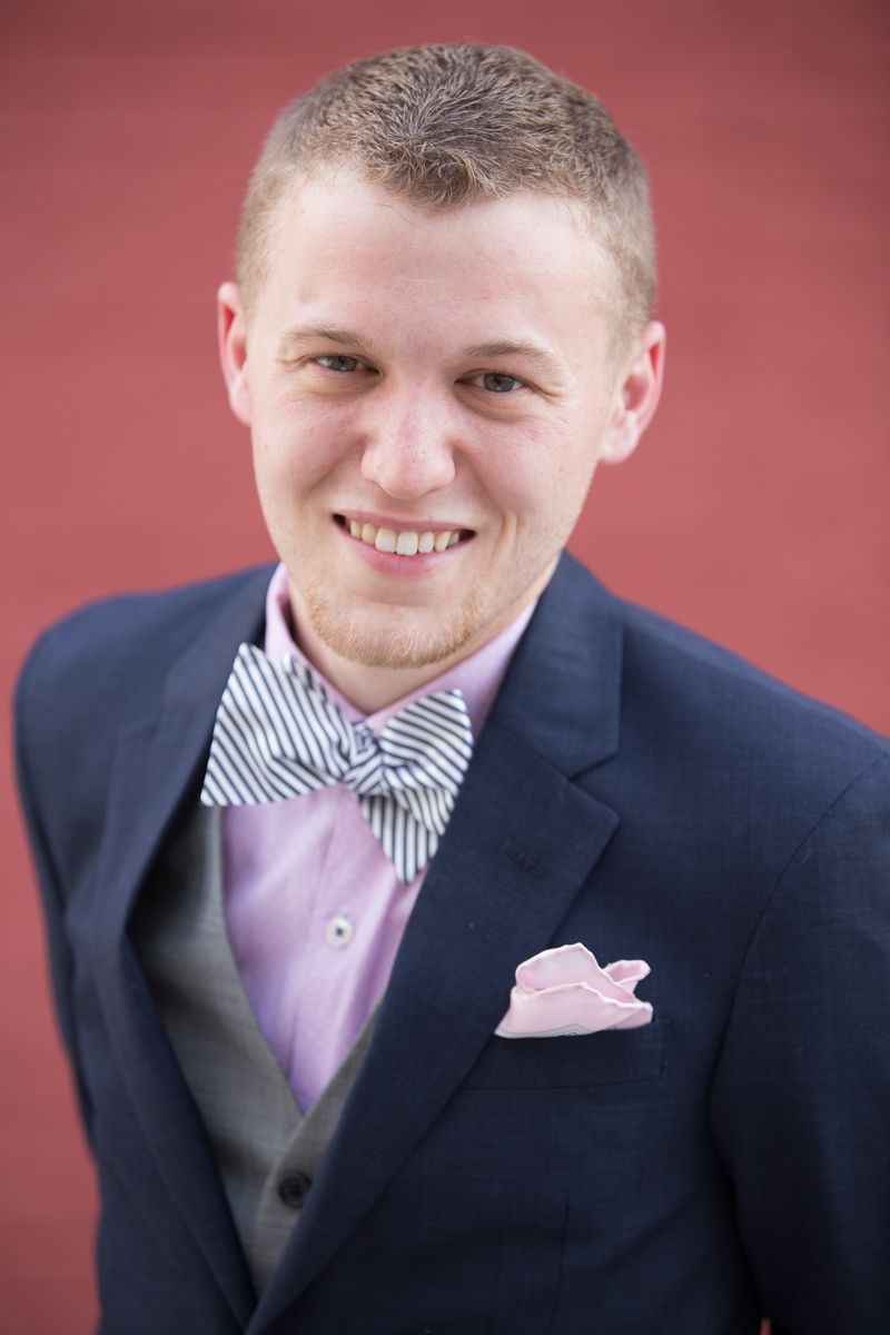 Professional portrait of someone in a navy suit with striped bow tie, pink pocket square and vest against red background.