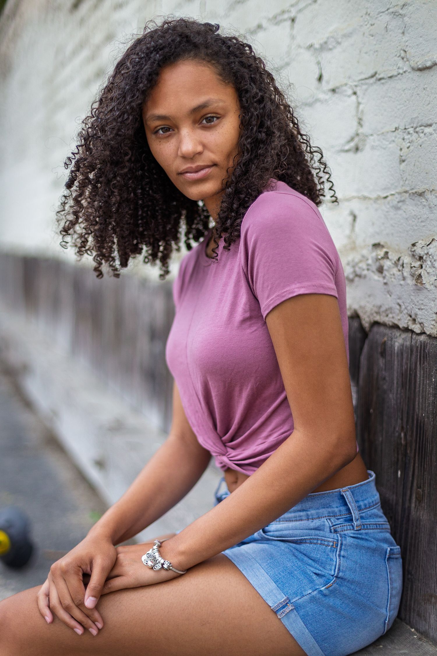 Person wearing purple t-shirt and blue denim shorts poses casually against white textured wall.