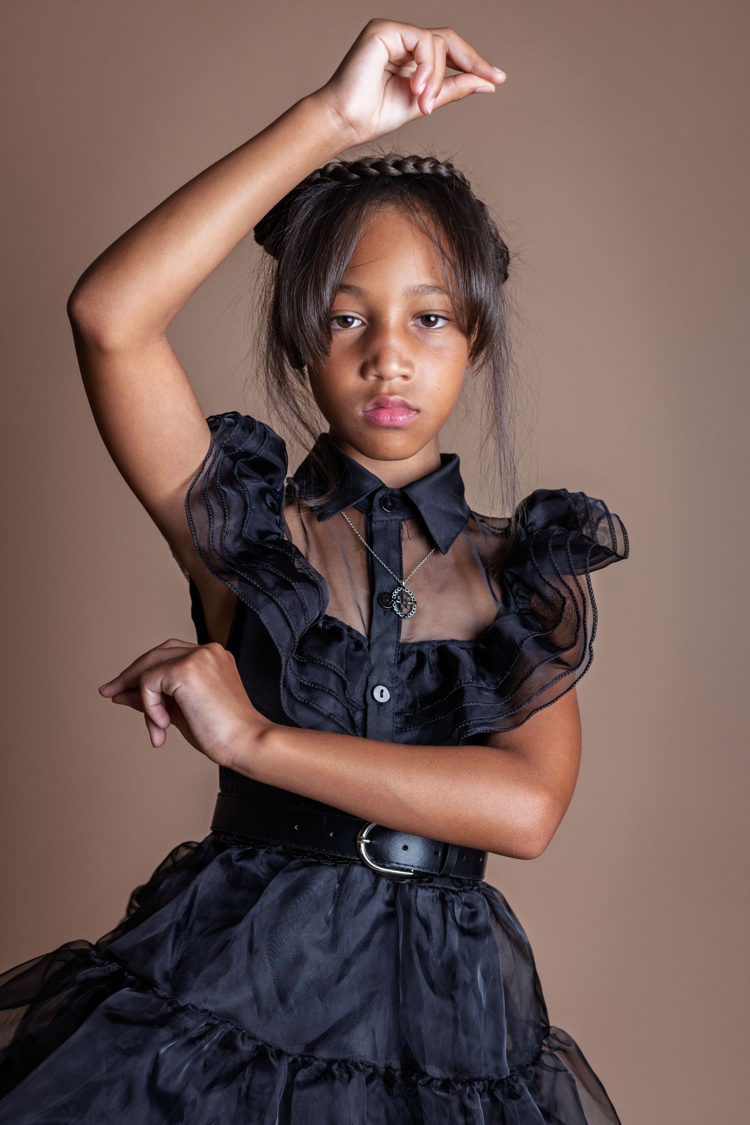 A young model in a black ruffled dress with a belt poses elegantly against a neutral brown background.