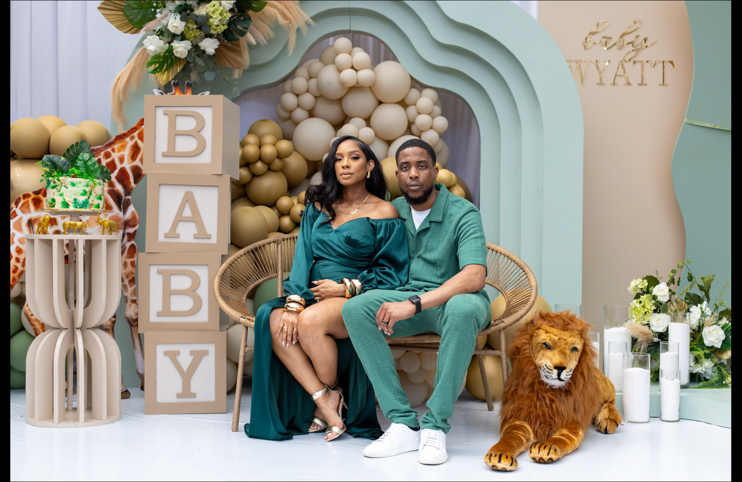 Couple in emerald green outfits pose at safari-themed baby shower with BABY blocks and lion plush.
