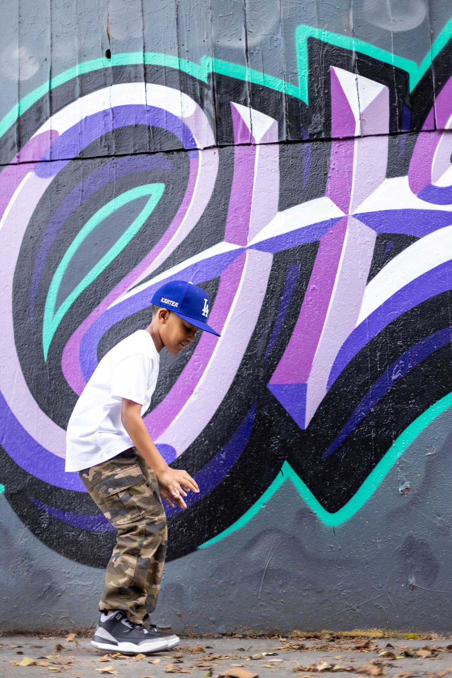 A person in camo pants and white shirt strikes a dance pose against a colorful graffiti wall with purple and turquoise designs.