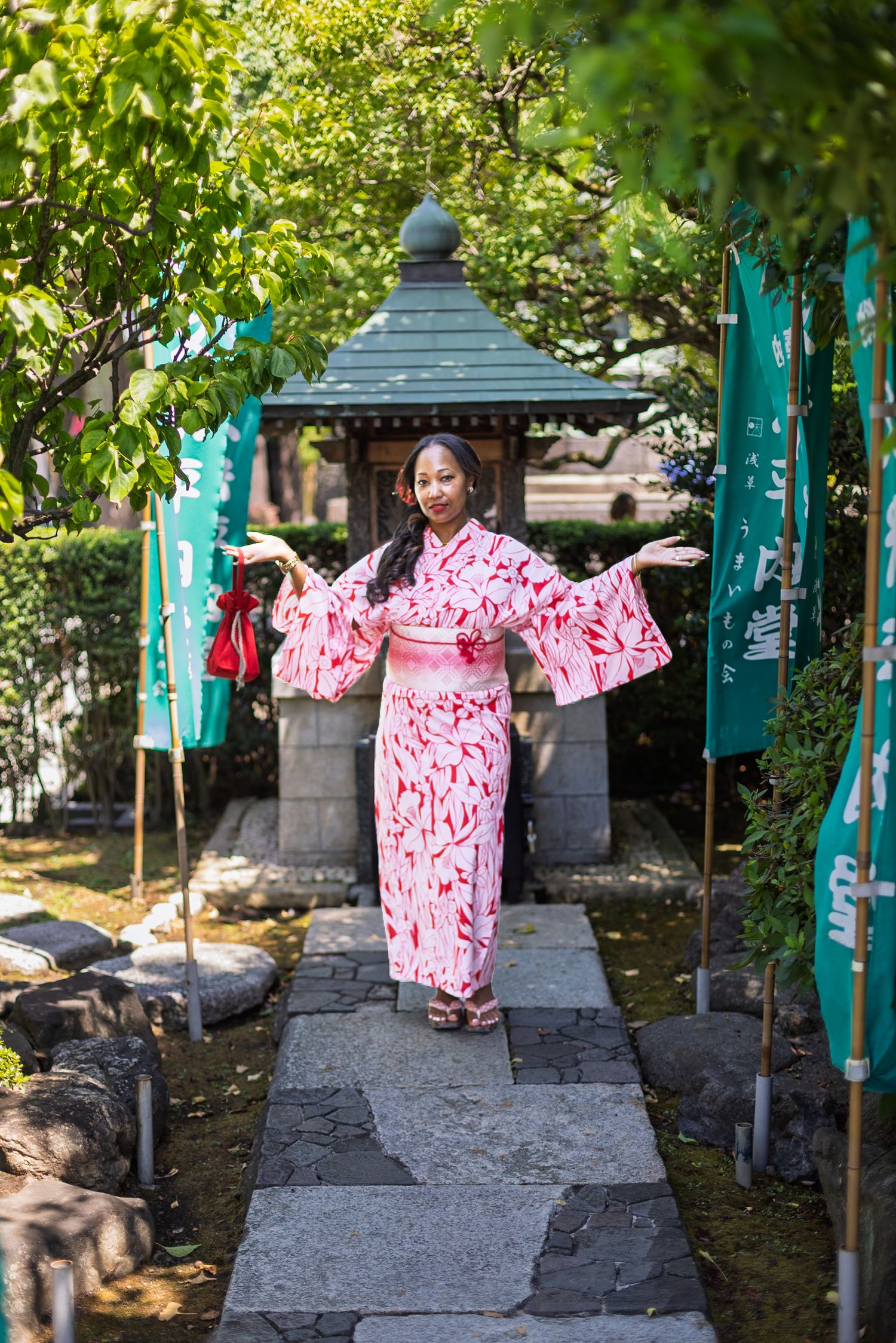 Woman in pink floral kimono poses on stone path in Japanese garden with green banners and stone lantern.