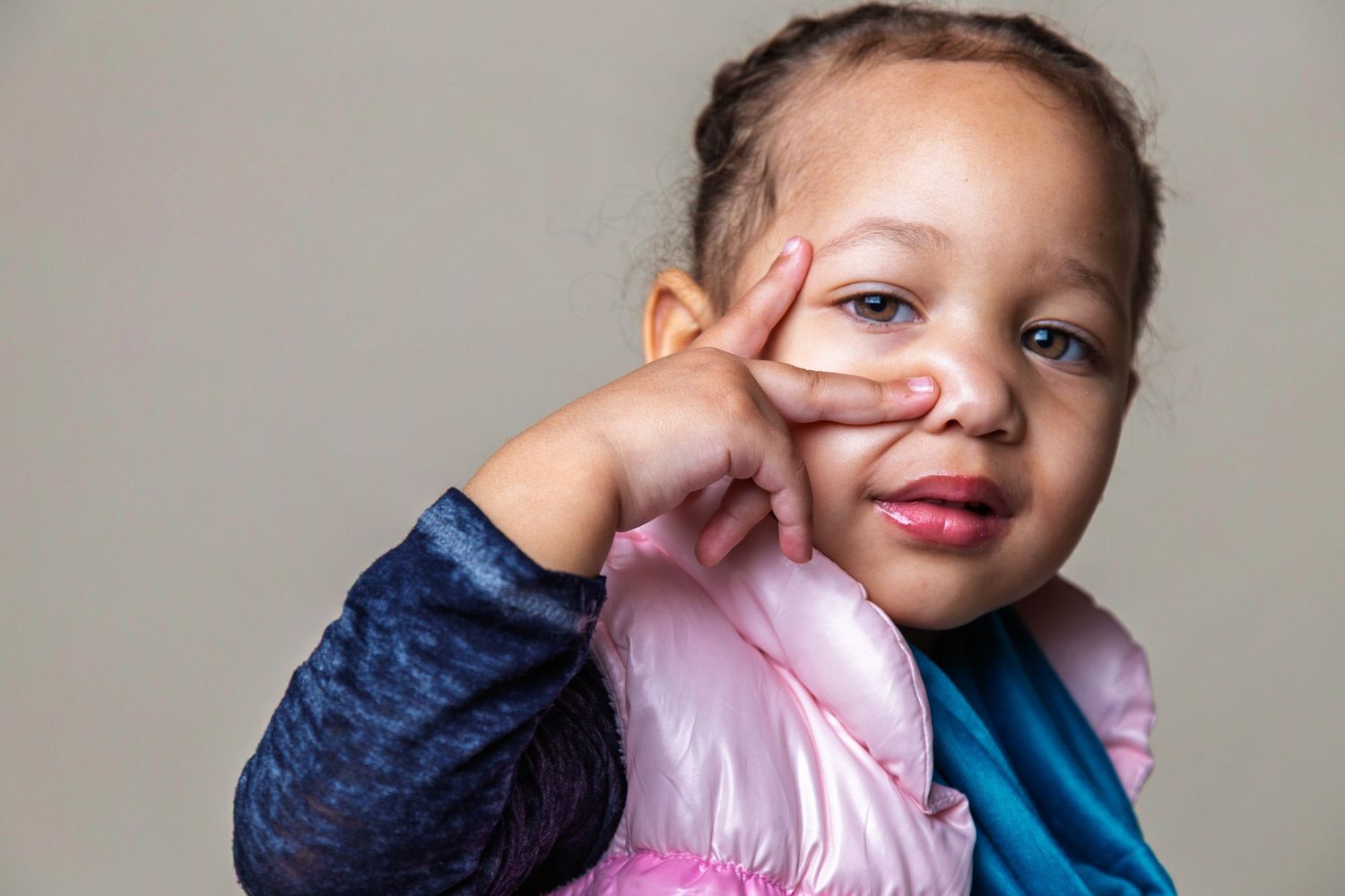 A young child in a blue and pink jacket poses with hand on cheek against a light background.
