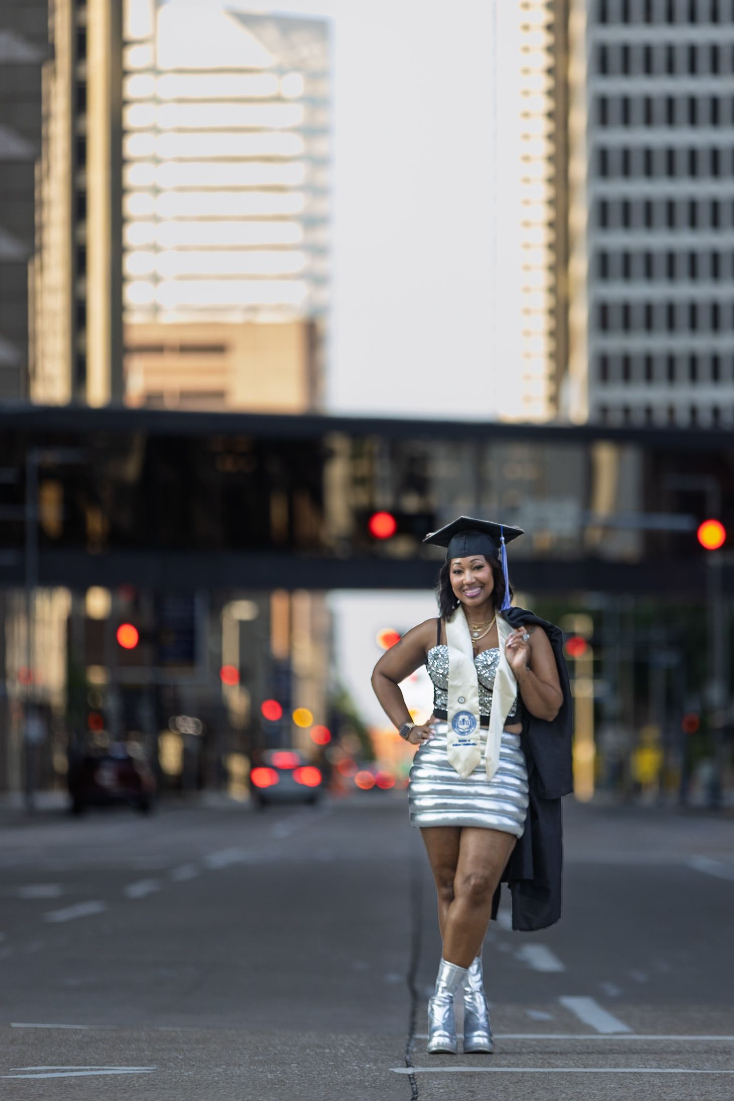 Graduate in silver dress and cap posing confidently on city street holding diploma in urban backdrop.
