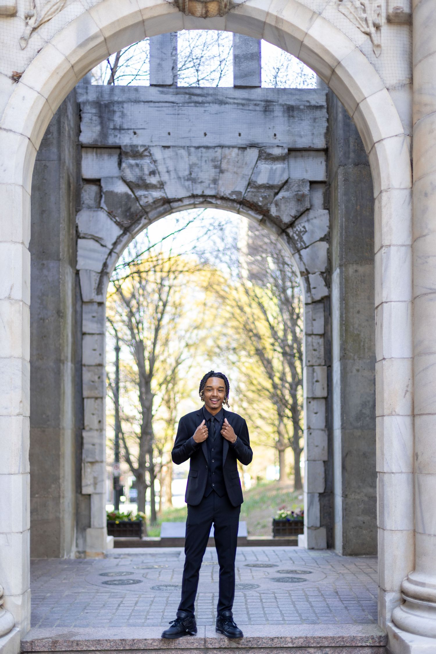 A person stands centered in a stone archway on a college campus during autumn with trees visible in the background.
