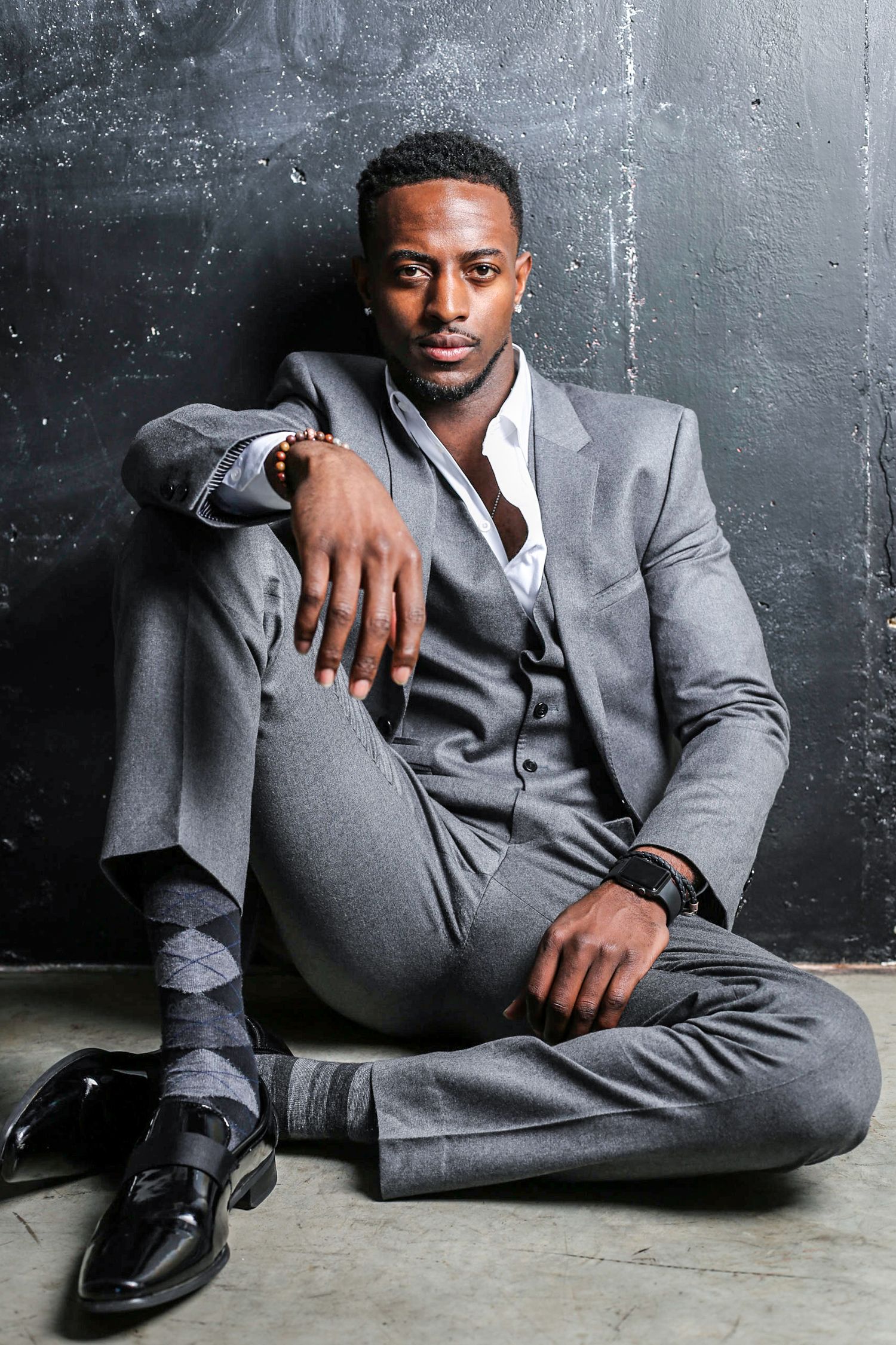 Professional portrait of a person in a well-fitted gray suit sitting casually against a dark textured wall.