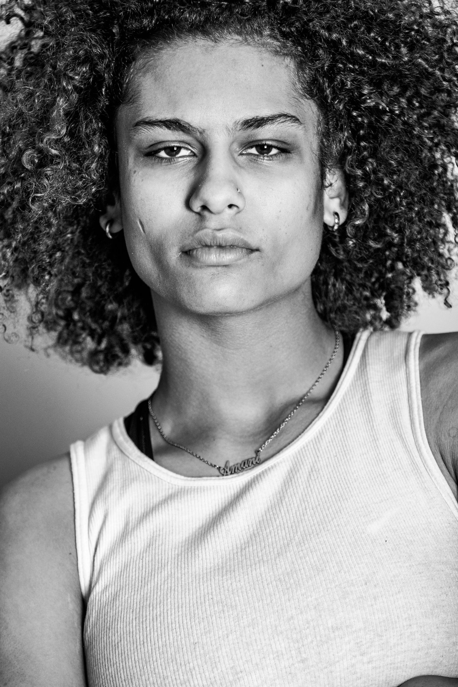 Black and white portrait of a young man with curly afro hair wearing a tank top and necklace, looking at camera.