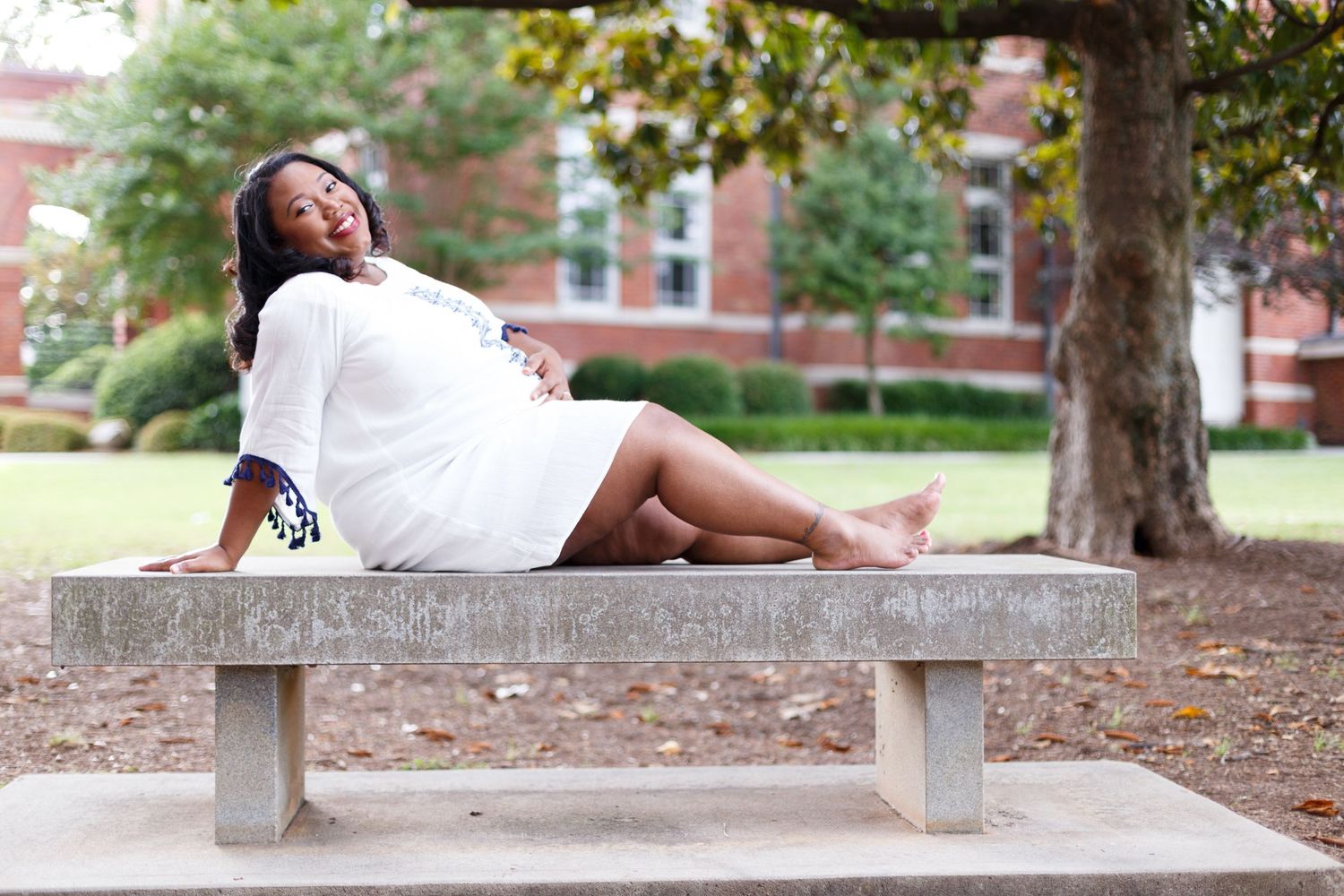 A person in a white dress relaxes on a stone bench in a scenic outdoor campus setting during a sunny day.
