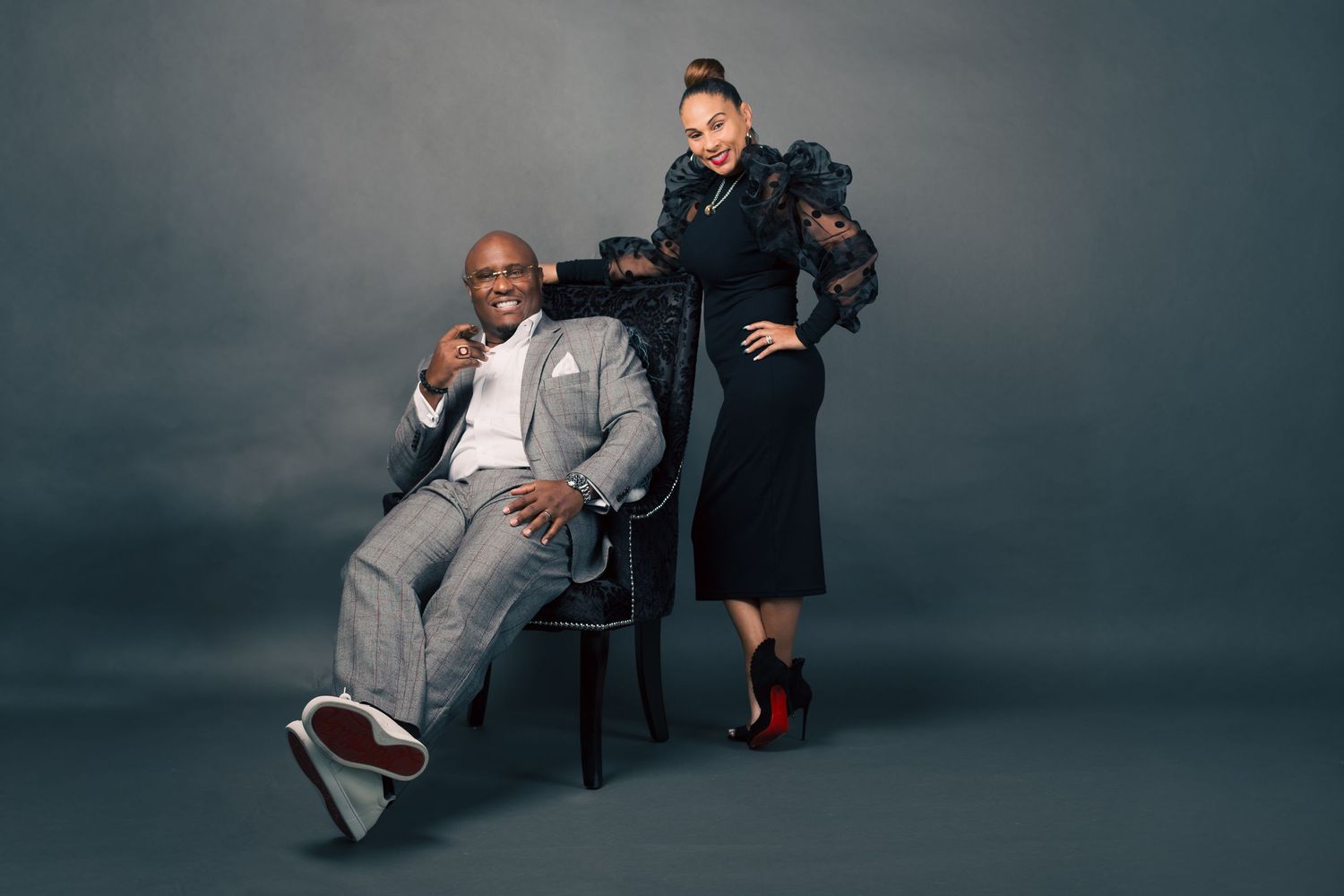 A stylish studio portrait of two people posing together, one seated in a chair wearing gray and one standing in black.