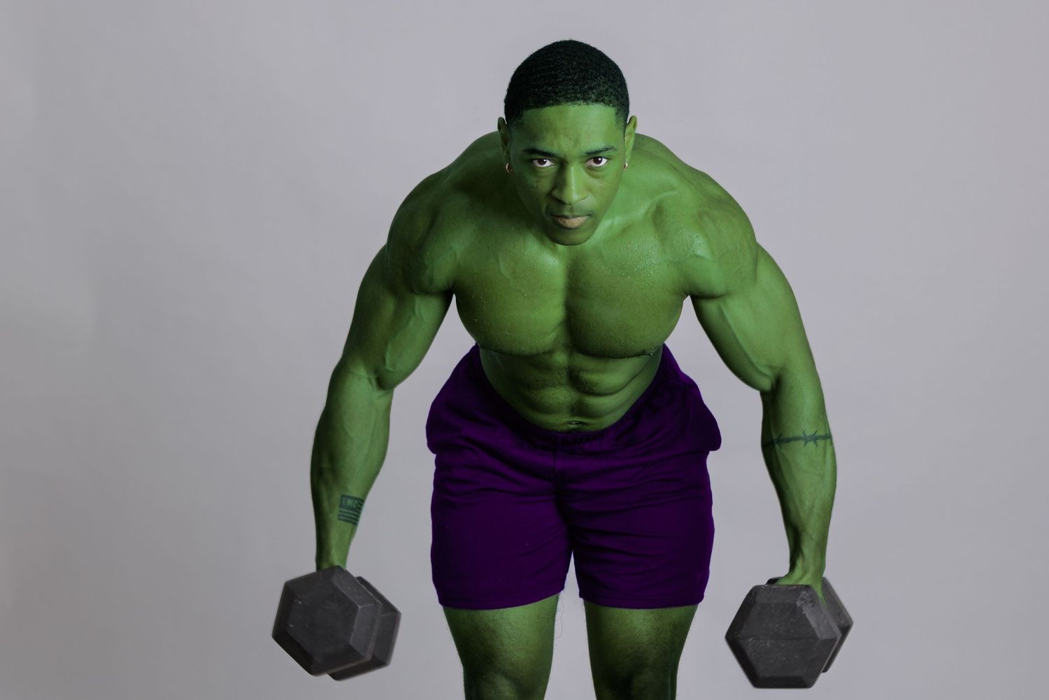 Muscular man painted green as the Hulk, holding dumbbells in a powerful pose against a gray background.
