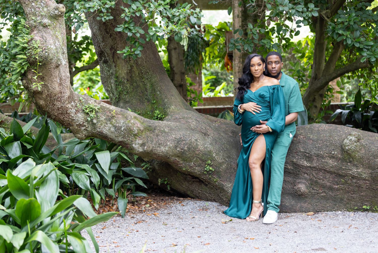 A couple in coordinating teal outfits poses together on a large tree branch in a lush garden setting.
