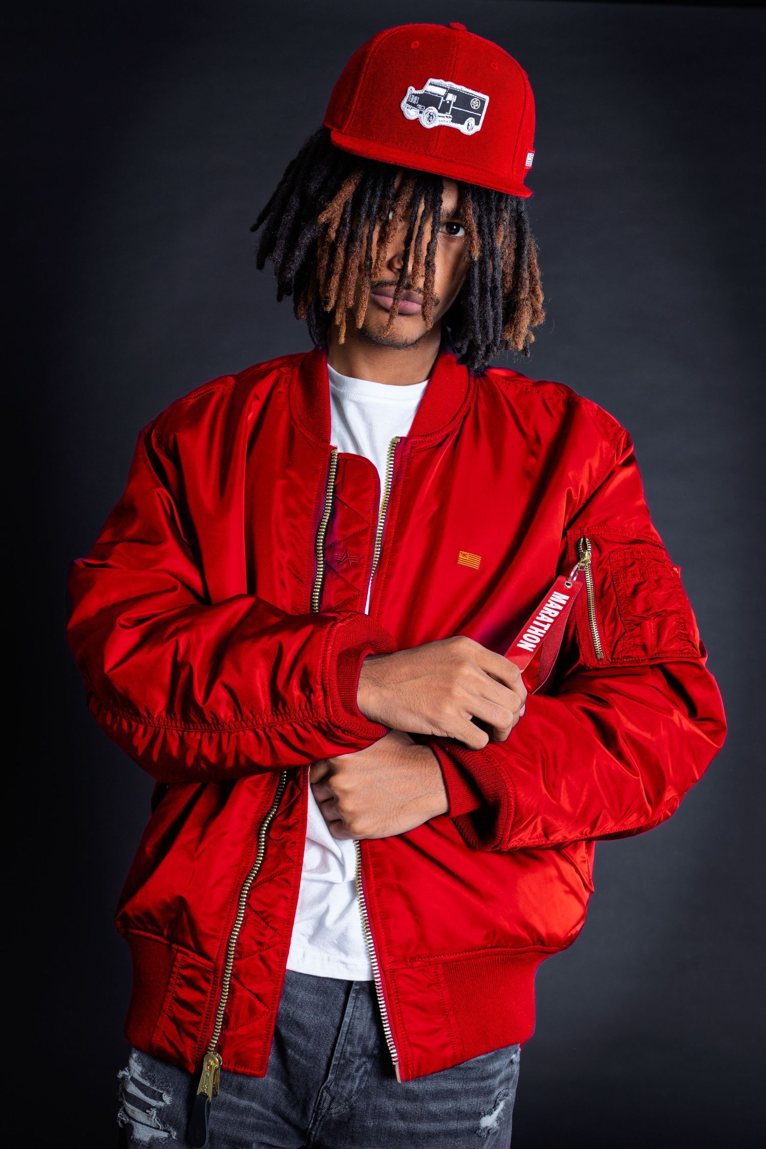 Young man with dreadlocks wearing a red bomber jacket and matching red cap poses confidently against dark background.