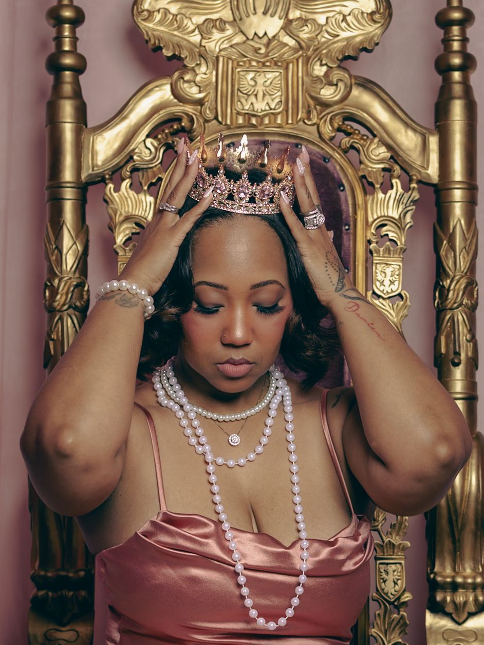 Person wearing a dusty rose satin dress and pearl necklaces poses against an ornate gold baroque backdrop.