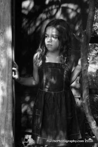 A black and white portrait of someone in a dark dress standing among trees and wooden posts in a moody setting.