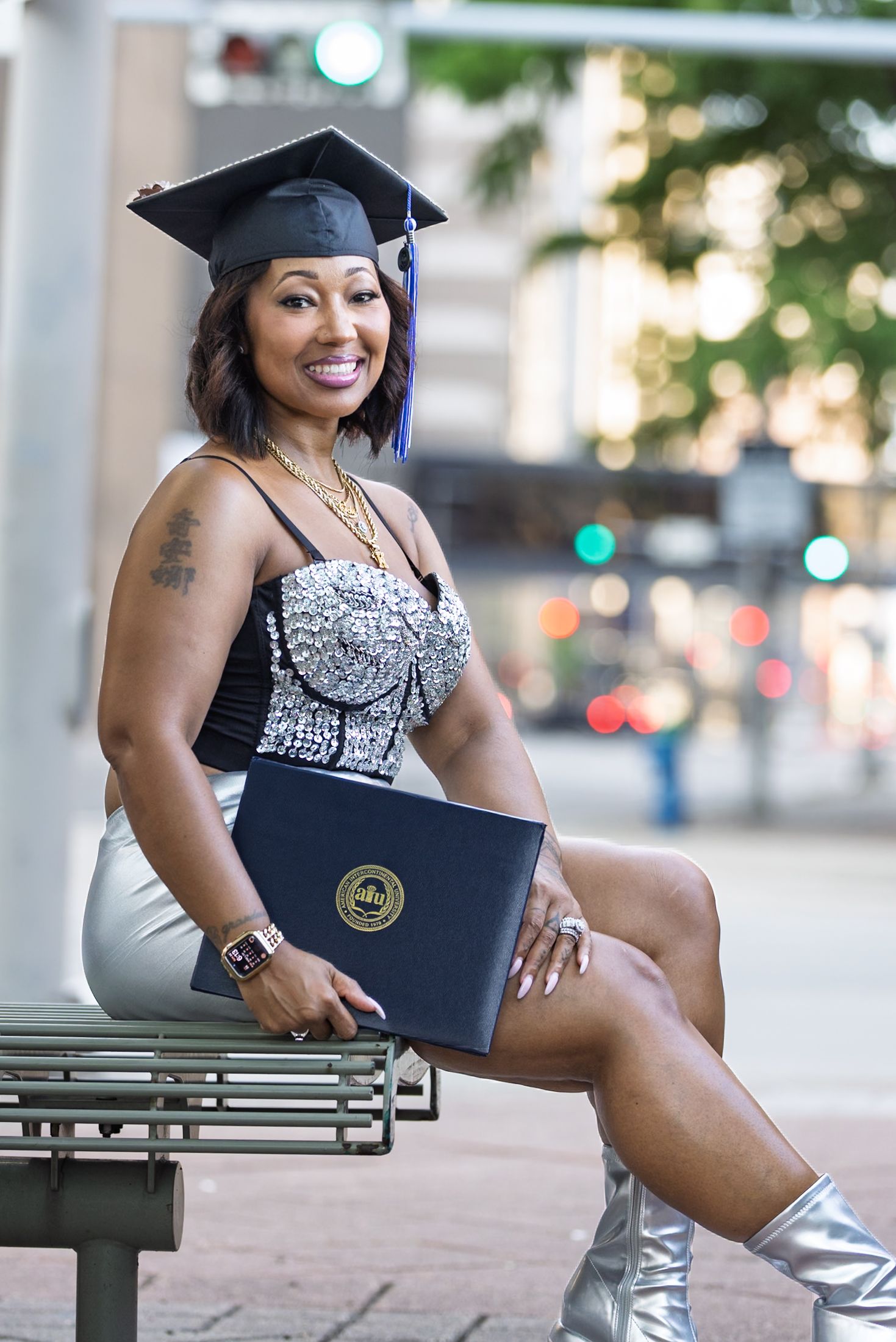 Smiling graduate in cap holding diploma on bench in sparkly outfit and silver boots in urban setting.