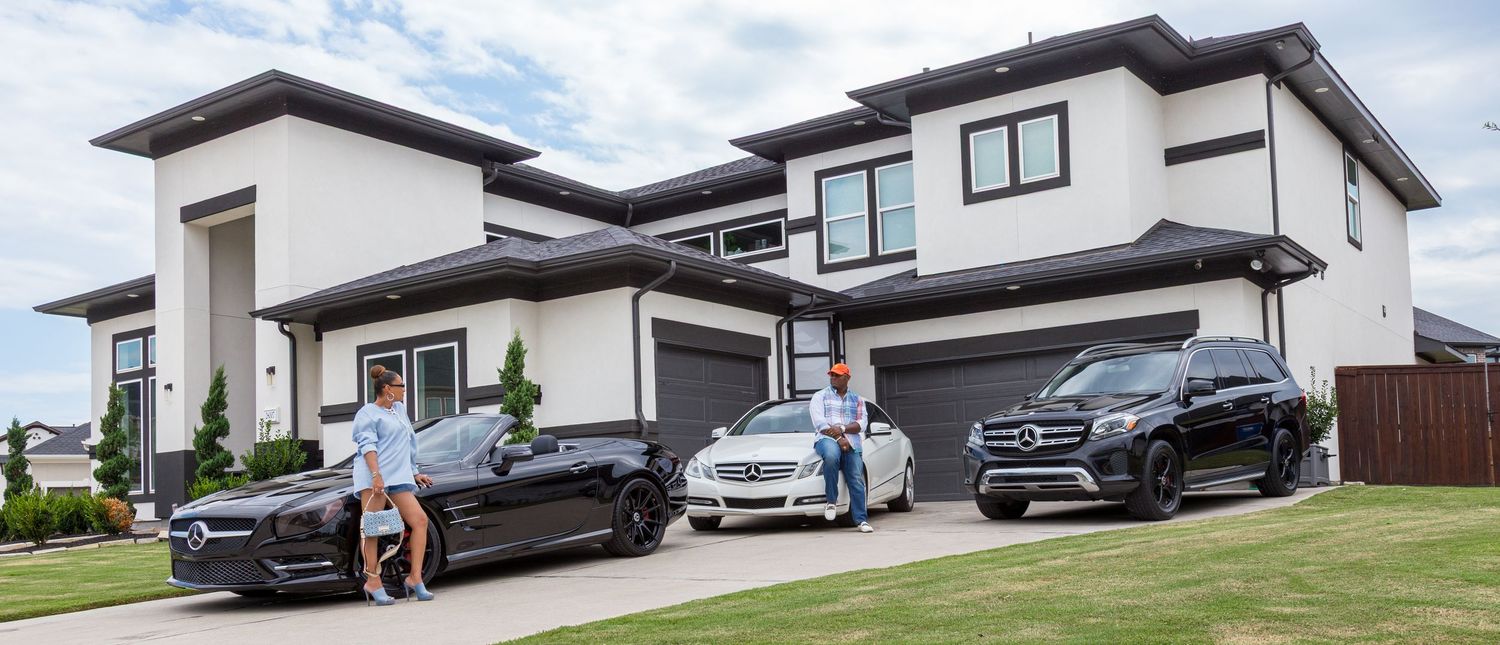 Two men pose with luxury cars including a Mercedes-Benz SUV and sedan in front of a modern two-story home.