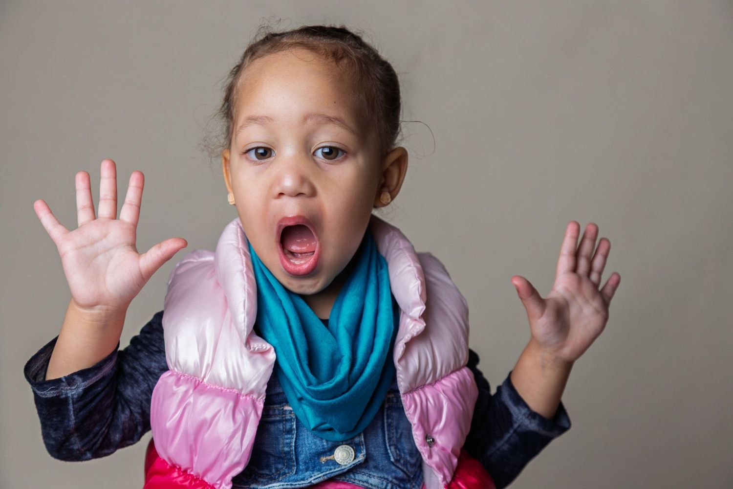 A child wearing a pink vest and blue scarf makes an excited expression with hands raised against a gray background.