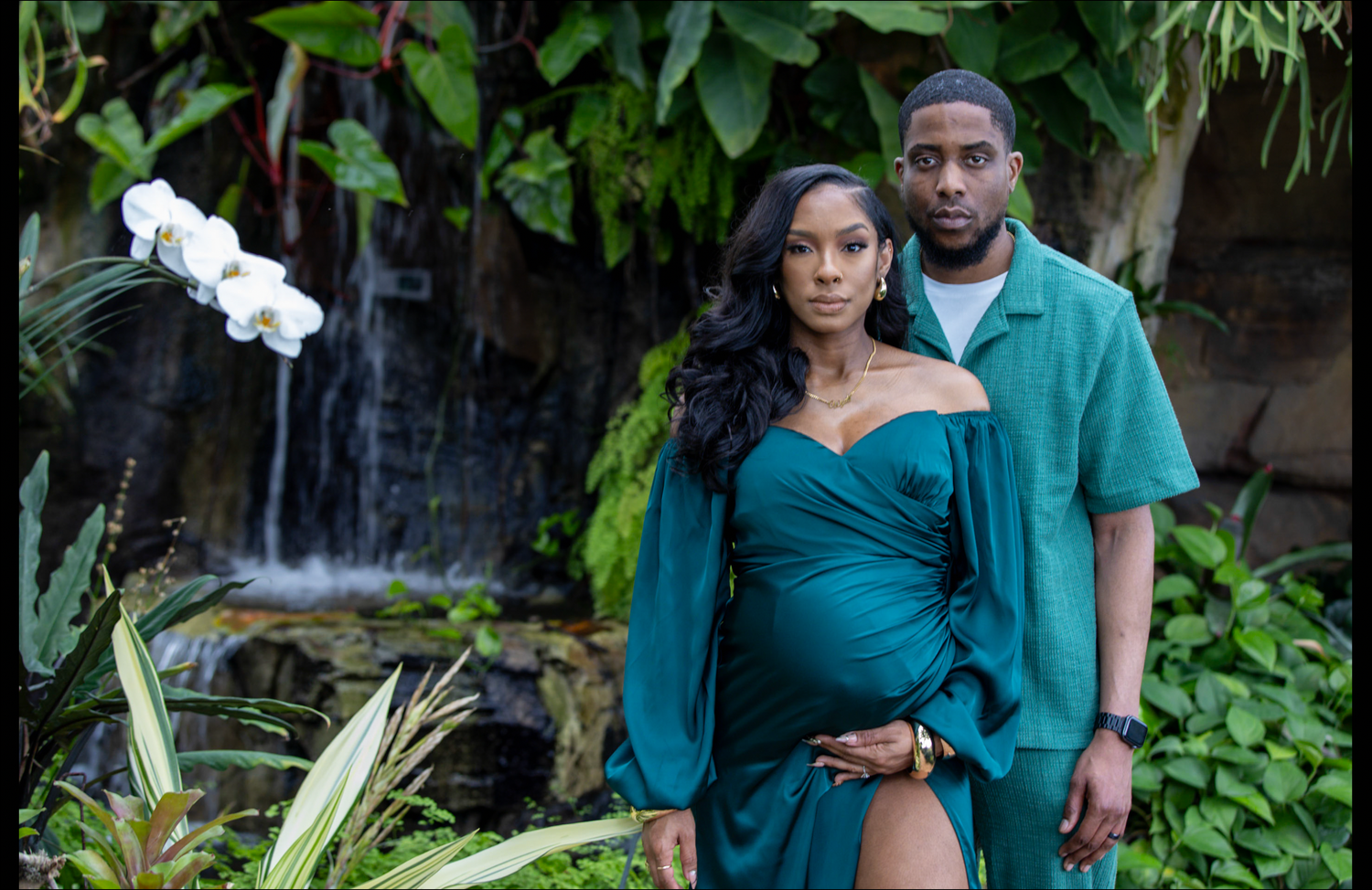 Pregnant woman in teal gown poses with partner in lush tropical garden with waterfall backdrop.