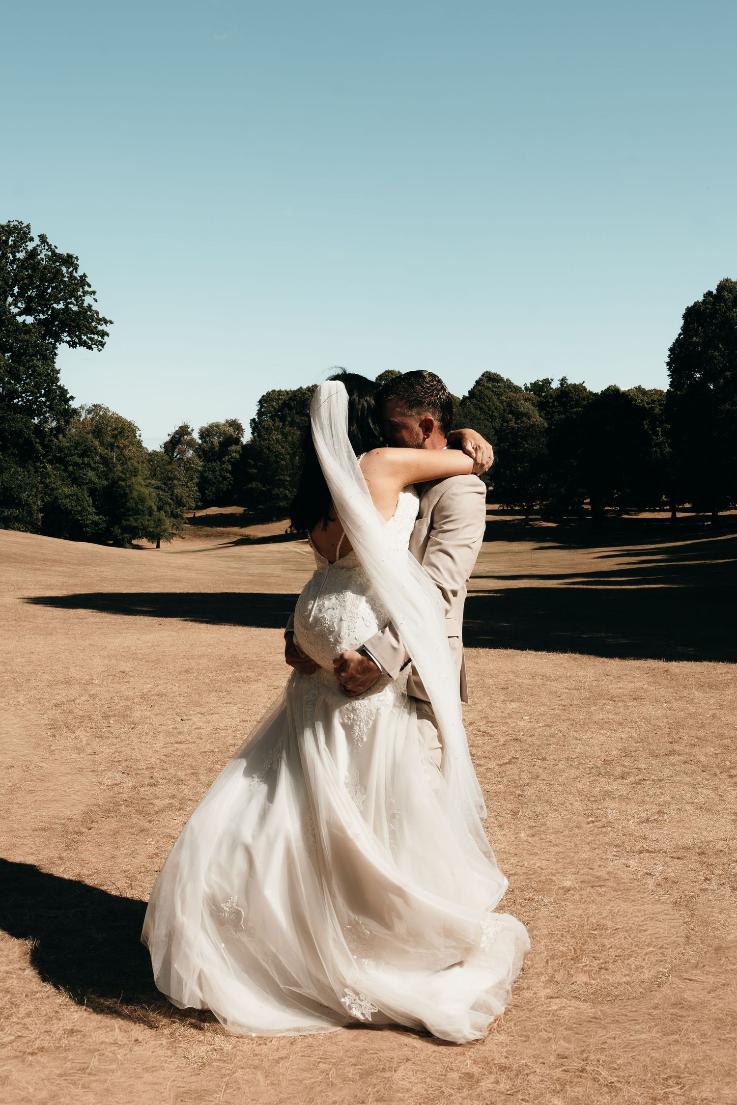 Bride and groom embracing in an outdoor park setting, bride in white gown with veil, groom in beige suit.