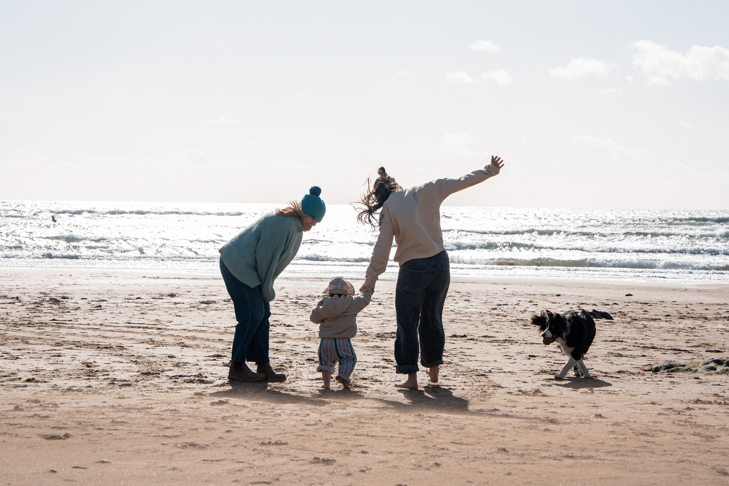 Two adults and a toddler playing with a dog on a sunny sandy beach near the ocean waves.