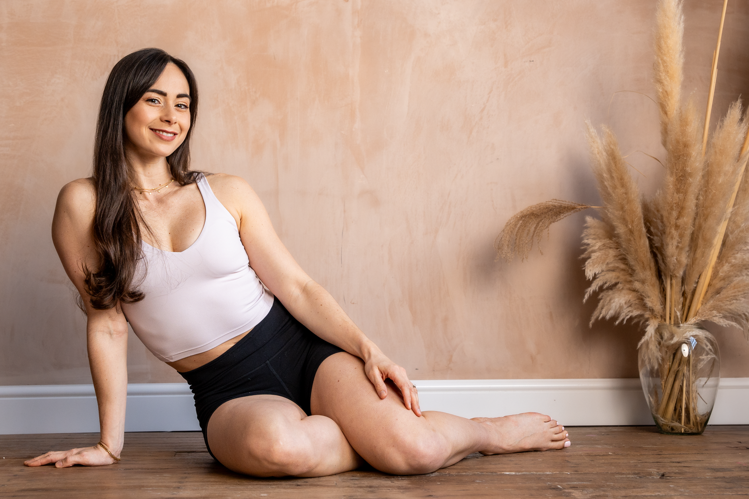 Smiling brunette woman in white crop top and black shorts sitting on floor near pampas grass.