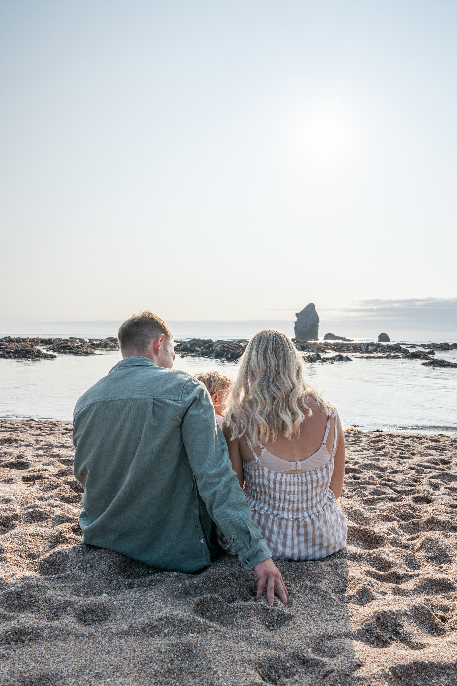 Couple sitting together on sandy beach from behind, gazing at calm ocean and rocky horizon at sunset.