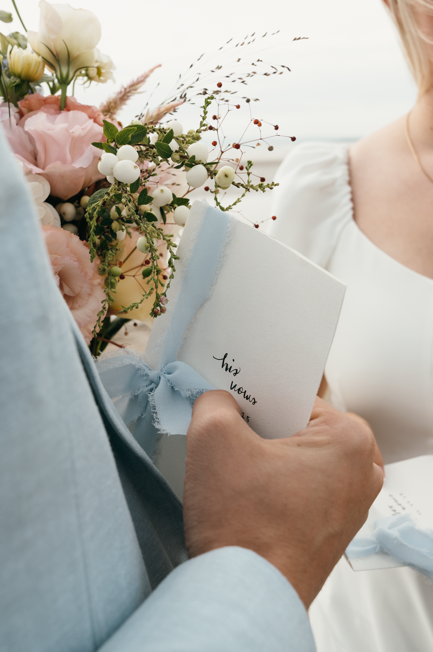 Groom in light blue suit holds his wedding vows booklet beside bride in white dress holding a pastel floral bouquet.