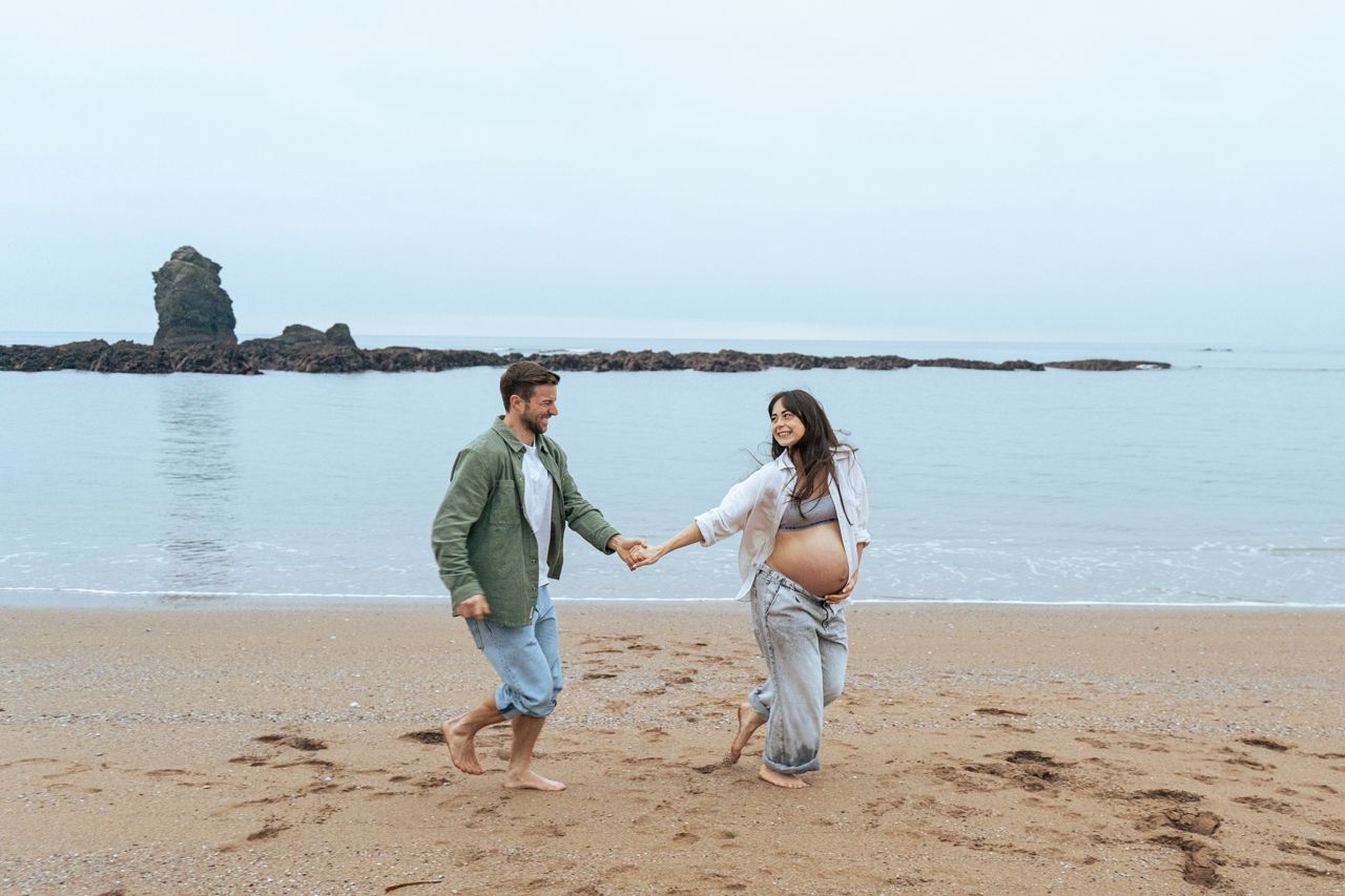 Happy pregnant couple holding hands and walking along sandy beach with rocky ocean coastline behind them.