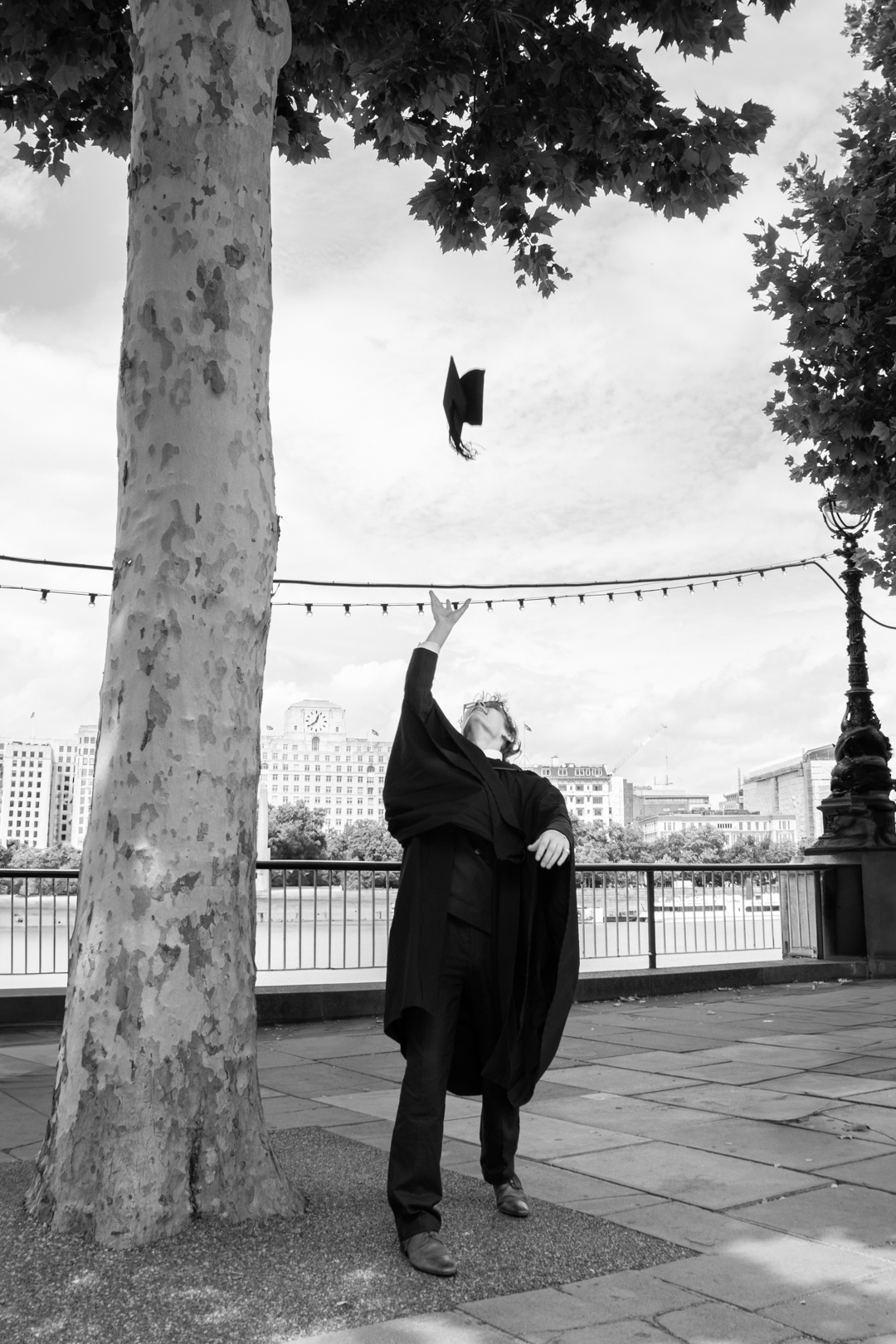A graduate in black gown tosses their graduation cap into the air beside a tree in black and white photo.