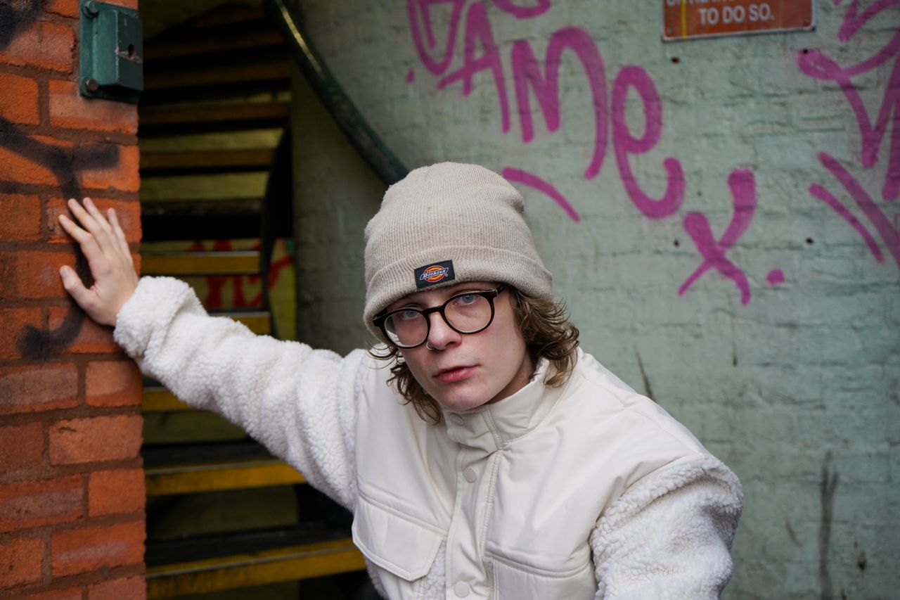 Person in white fleece jacket and beanie leaning against brick wall with pink graffiti art.