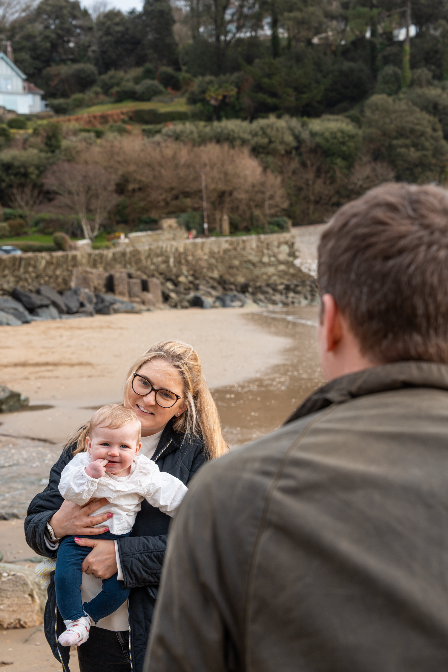 Mother holding smiling baby facing father on a sandy beach.