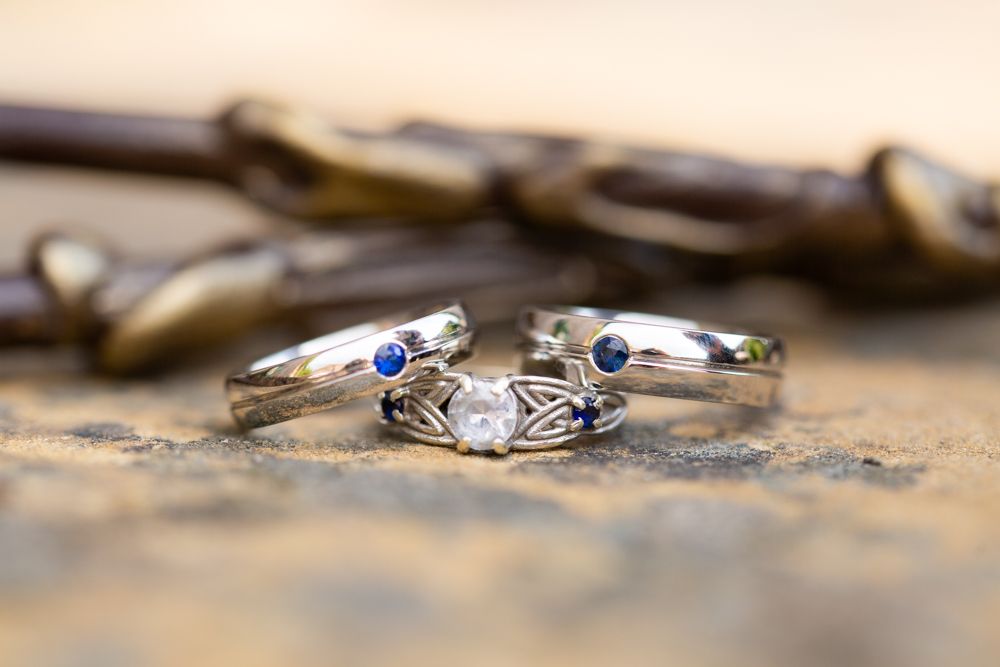 Three silver wedding rings with blue sapphires and a diamond centerpiece displayed on a stone surface.