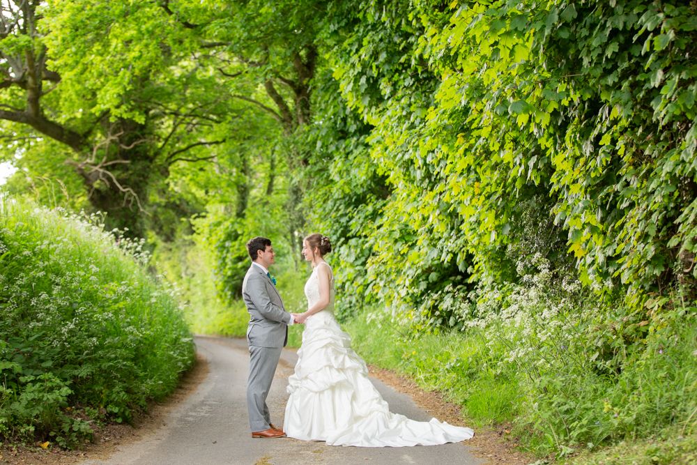 Bride and groom stand facing each other on a scenic tree-lined country lane in wedding attire.