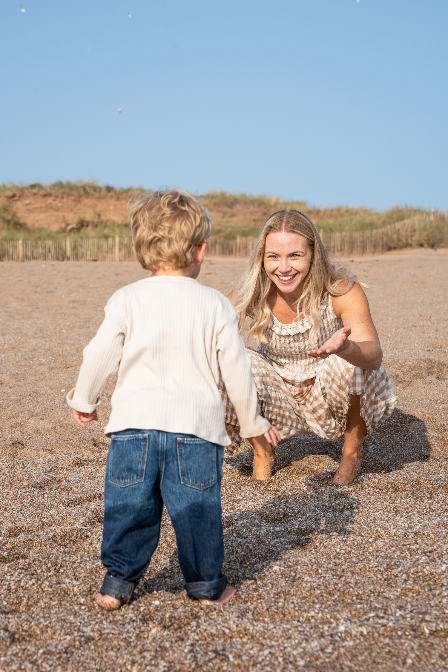 Smiling blonde mother crouching on pebbly beach reaching out to her young toddler on a sunny day.