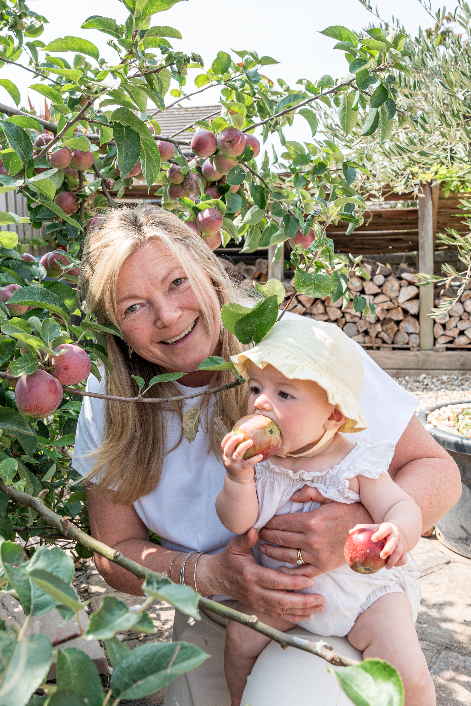 Grandmother holding baby eating apple under fruit tree in garden.