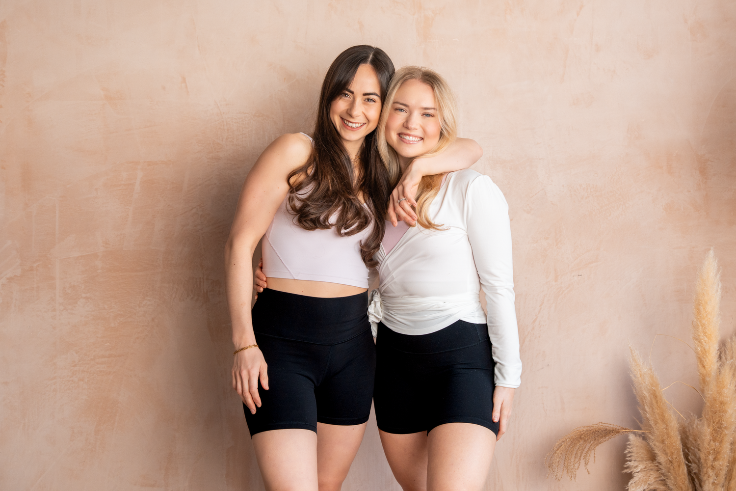 Two smiling women in matching black bike shorts pose together against a beige wall, wearing white crop tops.
