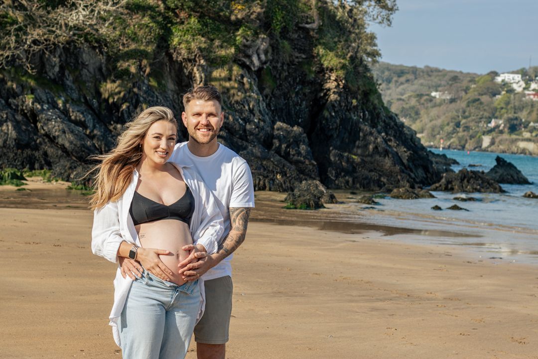 Smiling couple cradling pregnant belly on sunny sandy beach.