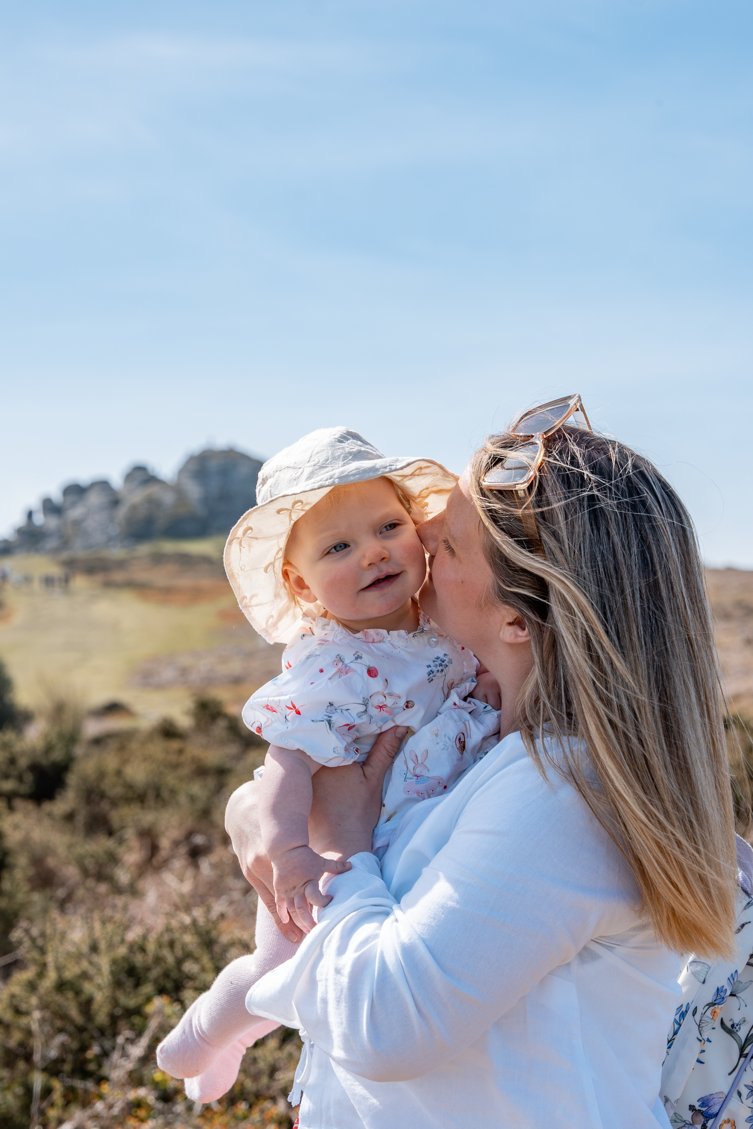 A mother kissing her smiling baby girl wearing a sun hat outdoors on a sunny day.