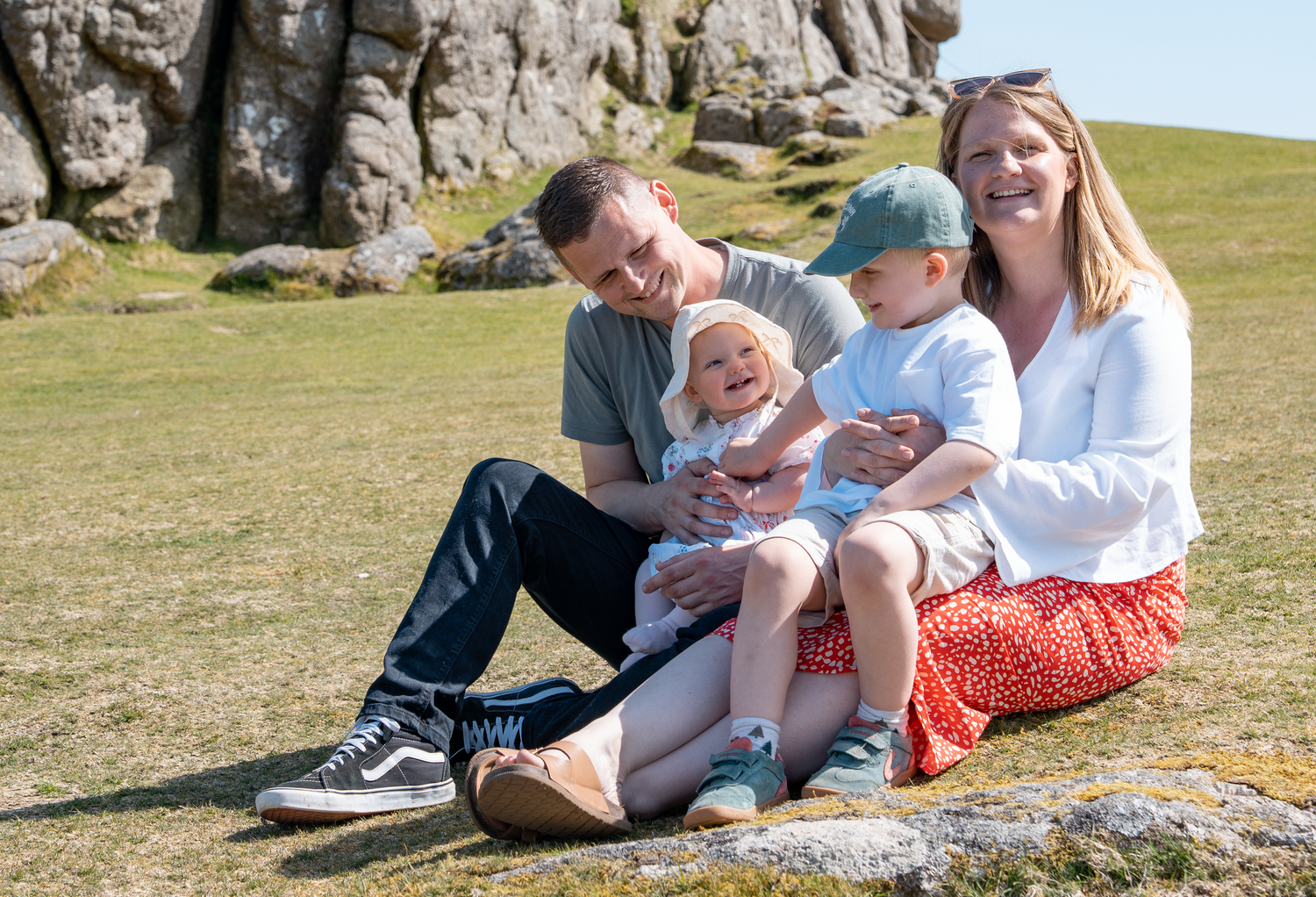 Happy family of four sitting on rocks in a sunny field, parents smiling with two young children on their laps.