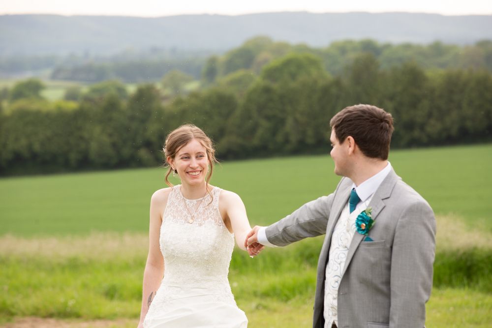 Smiling bride in lace gown holding grooms hand outdoors with green countryside landscape in background.