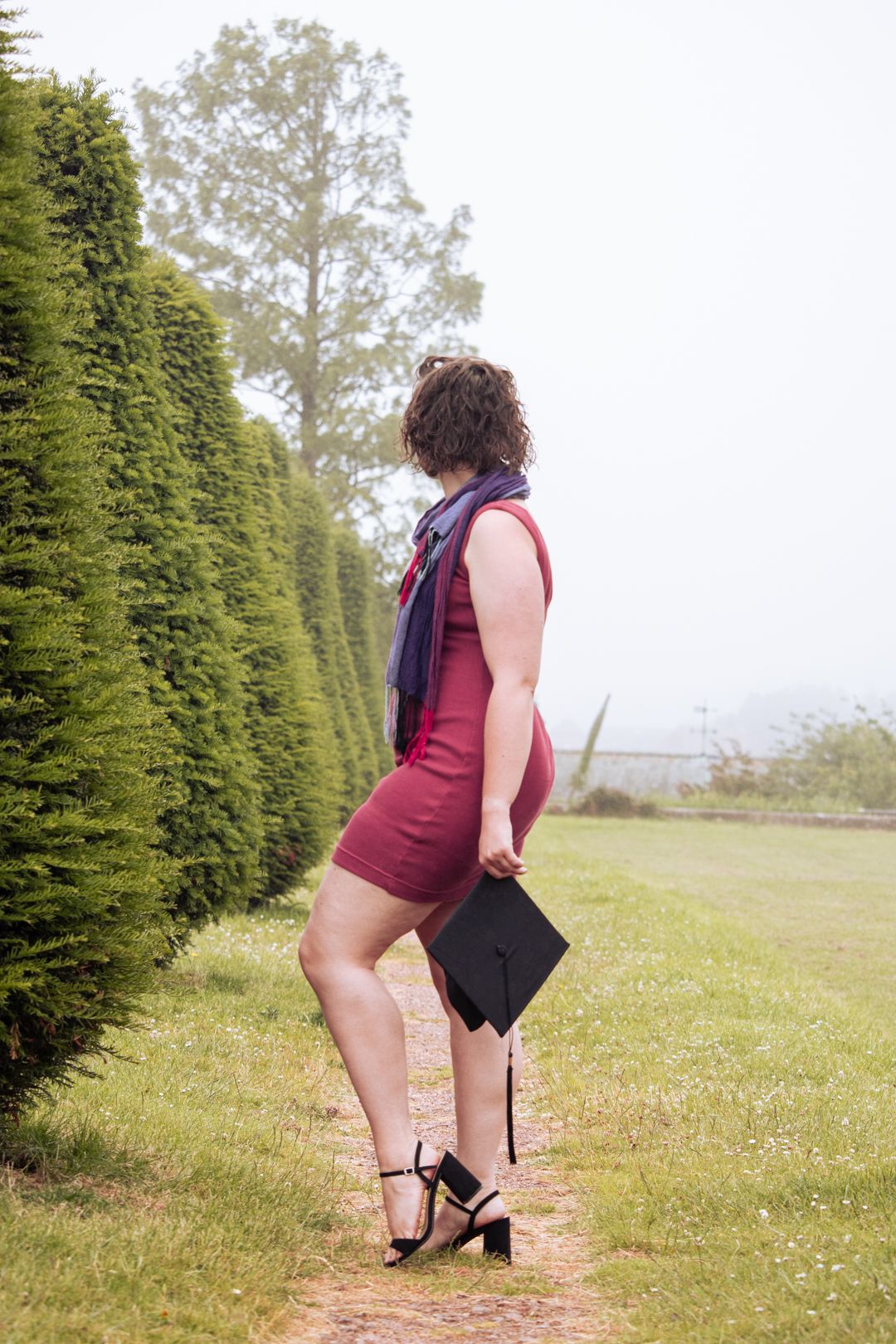 Young woman in burgundy dress holding graduation cap walks along garden path lined with tall hedges.